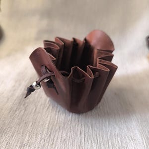 K&ouml;nnte beinhalten: Kleiner, brauner Lederbeutel mit Kordelzugverschluss. Der Beutel hat ein ger&uuml;schtes Design am oberen Rand und eine silberne Perle am Kordelzug. Das Leder hat eine satte, dunkle Farbe.