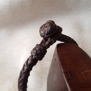 Puede incluir: Un brazalete de cuero marr&oacute;n con un dise&ntilde;o trenzado y un nudo en el extremo.