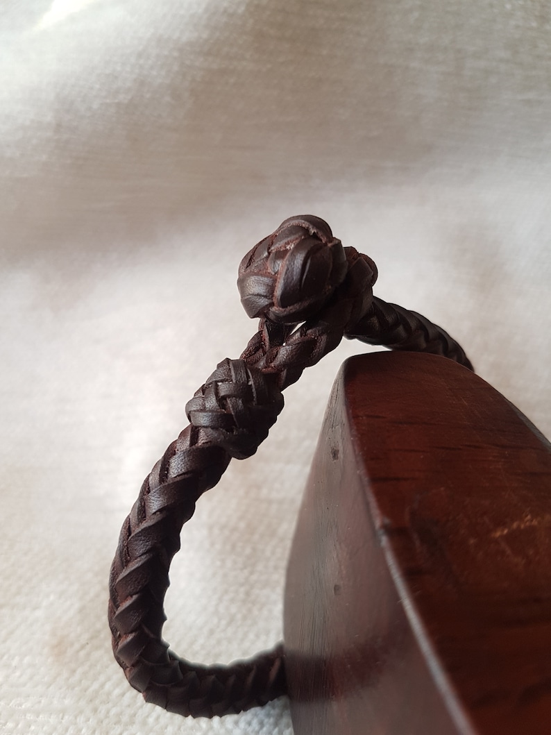 Puede incluir: Un brazalete de cuero marr&oacute;n con un dise&ntilde;o trenzado y un nudo en el extremo.