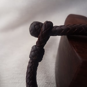 Puede incluir: Primer plano de una pulsera de cuero marr&oacute;n oscuro con un dise&ntilde;o trenzado. La pulsera est&aacute; atada en un nudo y descansa sobre una superficie de madera.
