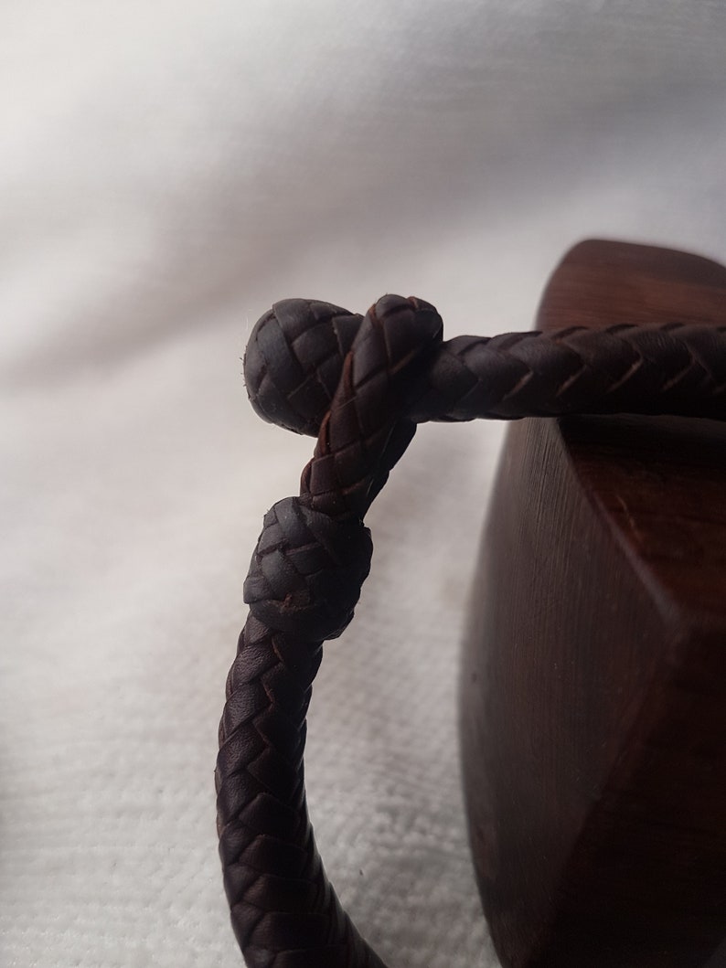 Puede incluir: Primer plano de una pulsera de cuero marr&oacute;n oscuro con un dise&ntilde;o trenzado. La pulsera est&aacute; atada en un nudo y descansa sobre una superficie de madera.