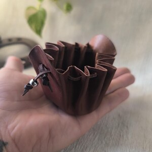 K&ouml;nnte beinhalten: Ein kleiner, brauner Lederbeutel mit Kordelverschluss und silbernem Perlenakzent. Der Beutel hat einen ger&uuml;schten Rand und wird in einer Hand gehalten. Das Leder hat eine satte, rotbraune Farbe.