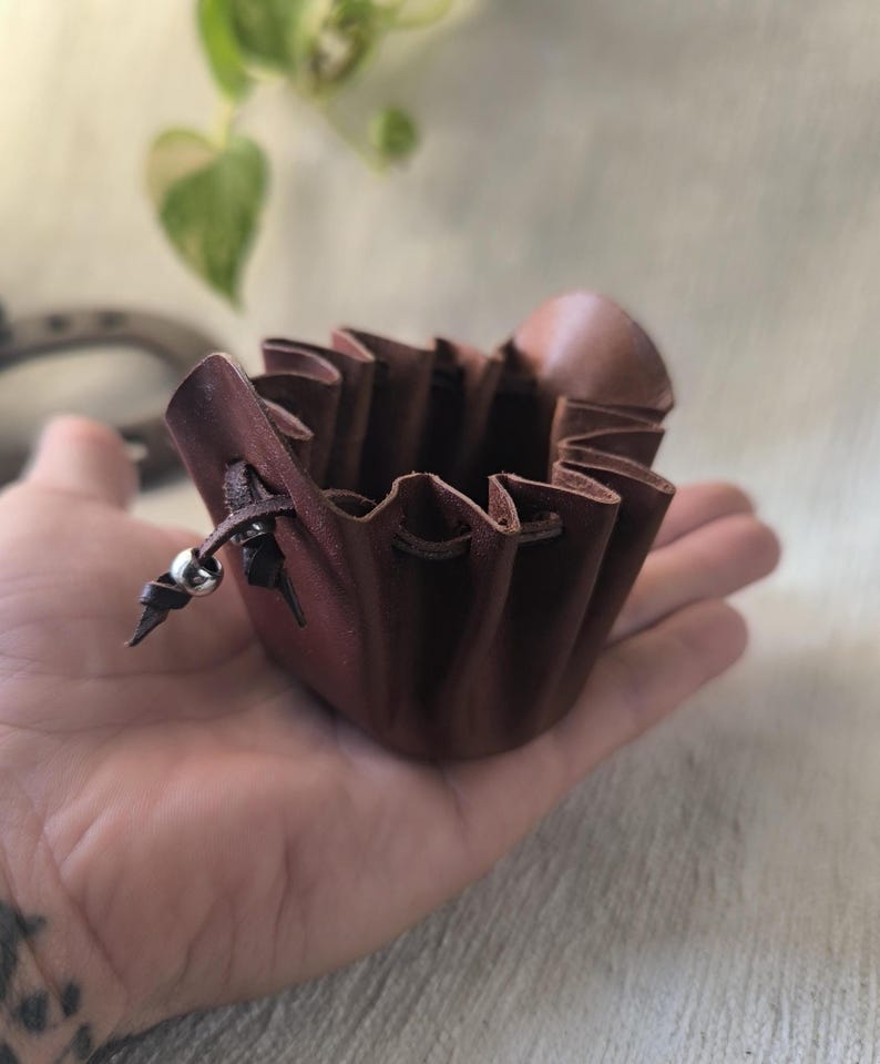 K&ouml;nnte beinhalten: Ein kleiner, brauner Lederbeutel mit Kordelverschluss und silbernem Perlenakzent. Der Beutel hat einen ger&uuml;schten Rand und wird in einer Hand gehalten. Das Leder hat eine satte, rotbraune Farbe.