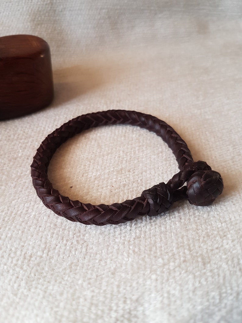 Puede incluir: Un brazalete de cuero marr&oacute;n oscuro con un dise&ntilde;o trenzado y un cierre anudado.