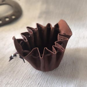 K&ouml;nnte beinhalten: Ein kleiner, brauner Lederbeutel mit Kordelzug. Der Beutel hat einen ger&uuml;schten oberen Rand und einen silberfarbenen Metallverschluss. Das Leder hat eine satte, dunkelbraune Farbe. Der Beutel ist f&uuml;r kleine Gegenst&auml;nde konzipiert.