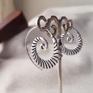 May include: A pair of silver-colored hoop earrings with a spiral design. The earrings are displayed on a silver earring stand. The spiral design features a series of parallel lines, creating a textured effect. The earrings have a hook closure.