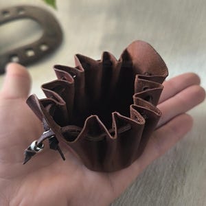 K&ouml;nnte beinhalten: Ein kleiner, dunkelbrauner Lederbeutel mit Kordelzug. Der Beutel hat einen ger&uuml;schten oberen Rand und einen silberfarbenen Metallakzent am Kordelzug. Der Beutel wird in der Hand einer Person gehalten.