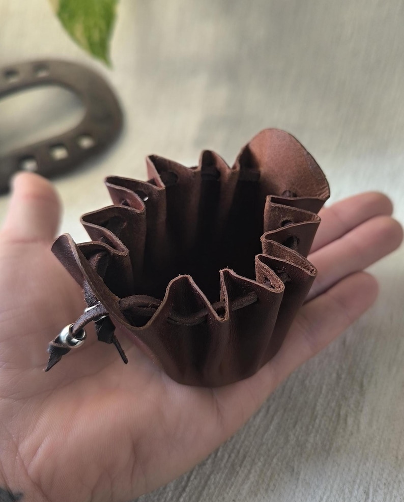 K&ouml;nnte beinhalten: Ein kleiner, dunkelbrauner Lederbeutel mit Kordelzug. Der Beutel hat einen ger&uuml;schten oberen Rand und einen silberfarbenen Metallakzent am Kordelzug. Der Beutel wird in der Hand einer Person gehalten.
