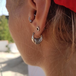 Puede incluir: Primer plano de la oreja de una persona con varios pendientes de plata. Los pendientes incluyen un pendiente de aro con un diseño decorativo, un pequeño pendiente de botón y un pendiente de aro con un pequeño círculo.