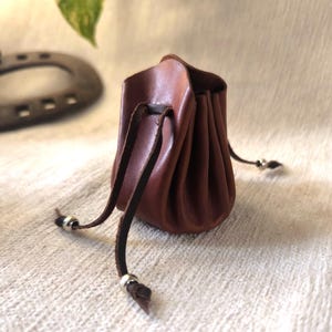 K&ouml;nnte beinhalten: Kleine, braune Ledertasche mit Kordelzug. Die Tasche ist oben gerafft und hat einen dunkelbraunen Lederkordelzug mit silberfarbenen Metallperlen an den Enden. Zum Tragen kleiner Gegenst&auml;nde.