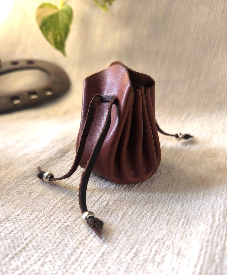 K&ouml;nnte beinhalten: Kleine, braune Ledertasche mit Kordelzug. Die Tasche ist oben gerafft und hat einen dunkelbraunen Lederkordelzug mit silberfarbenen Metallperlen an den Enden. Zum Tragen kleiner Gegenst&auml;nde.