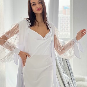 May include: A white dress with a sheer overlay featuring pearl embellishments on the sleeves. The dress has a cowl neck and a fitted silhouette.