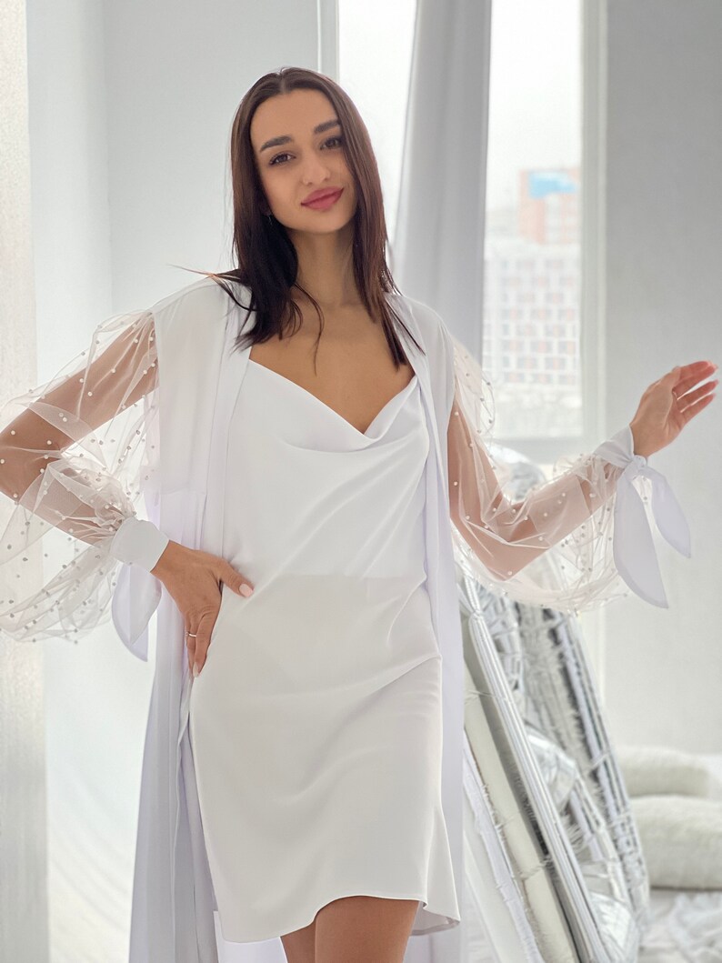 May include: A white dress with a sheer overlay featuring pearl embellishments on the sleeves. The dress has a cowl neck and a fitted silhouette.