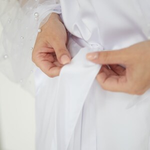 May include: A white satin robe with a sash tie and sheer sleeves with pearl embellishments.