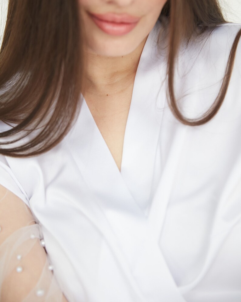 May include: A close-up of a woman's neckline wearing a white button-down shirt with a V-neck. The shirt has a sheer, beaded overlay on the sleeve.