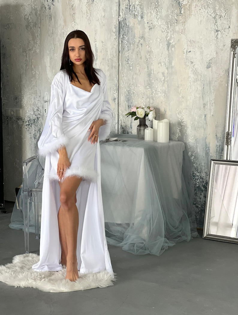 May include: A white satin robe with a long, flowing skirt and a shawl collar. The robe has a white feather trim around the cuffs and hem. The model is standing on a white fur rug.