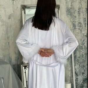 May include: A white satin robe with a fluffy white trim around the cuffs and waist. The robe is long and flowing, and the fabric is soft and luxurious.
