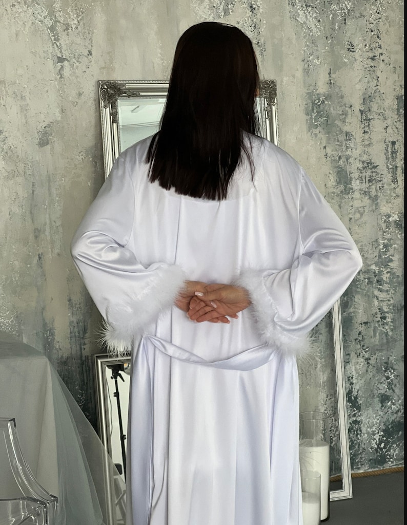 May include: A white satin robe with a fluffy white trim around the cuffs and waist. The robe is long and flowing, and the fabric is soft and luxurious.