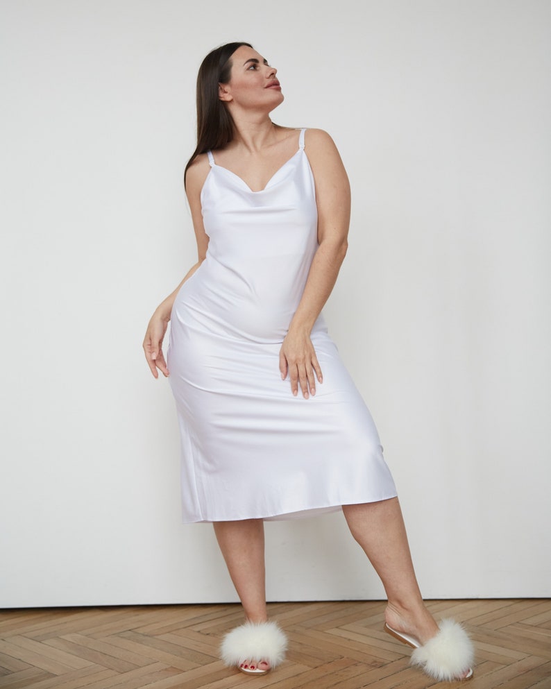 May include: A white slip dress with a cowl neck and thin straps. The dress is made of a silky material and falls to the mid-calf. The model is wearing white furry slippers.