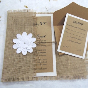 Puede incluir: Juego de invitaciones de boda rústicas con una envoltura de arpillera con una flor de papel blanca y cuerda. La invitación incluye los nombres "Brett" y la fecha "TH OF MAY, AD SIXTEEN". También se incluye una tarjeta RSVP y un sobre marrón.