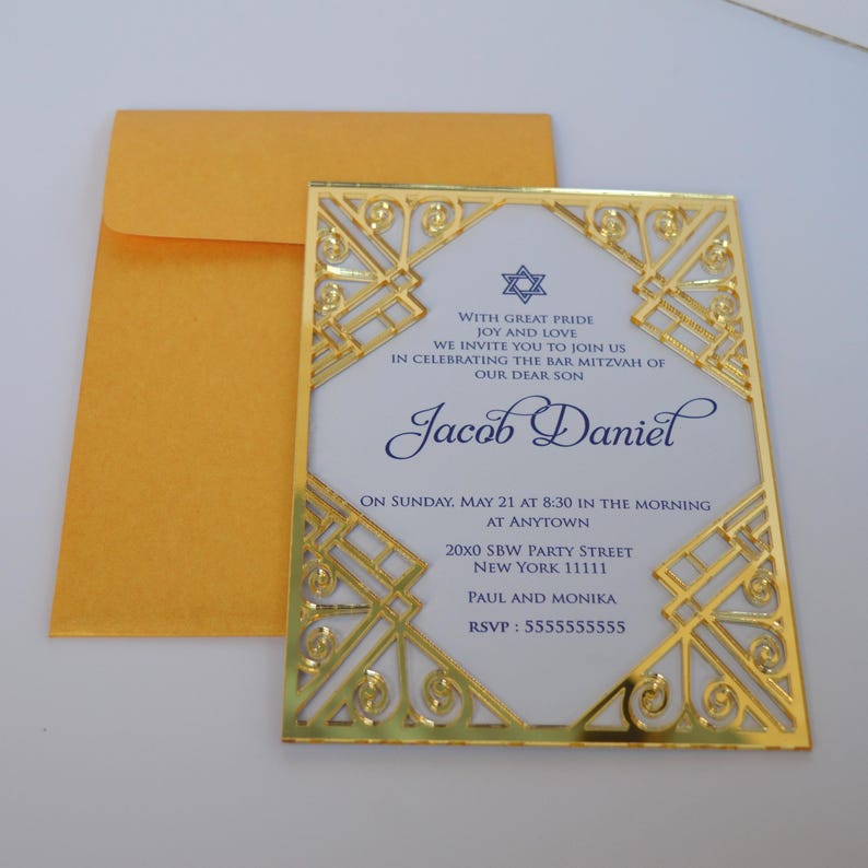 This is a beautiful, elegant Bar Mitzvah invitation featuring a striking gold laser-cut mirror acrylic frame with an intricate Art Deco style. The gold geometric swirls surround a white cardstock center printed with navy blue text and a Star of David at the top.

The combination of the formal serif typography for the details and the flowing script for Jacob's name creates a sophisticated and modern look.