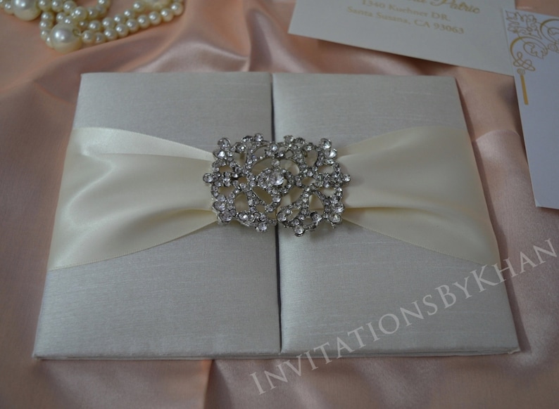 May include: A white satin invitation box with a large, ornate rhinestone embellishment. The box is closed and has a satin ribbon tied around it.