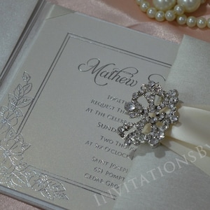 May include: A white wedding invitation with silver foil accents and a floral design. The invitation is tied with a white satin ribbon and a rhinestone brooch. The text on the invitation reads "Mathew and" and includes the wedding date and time.