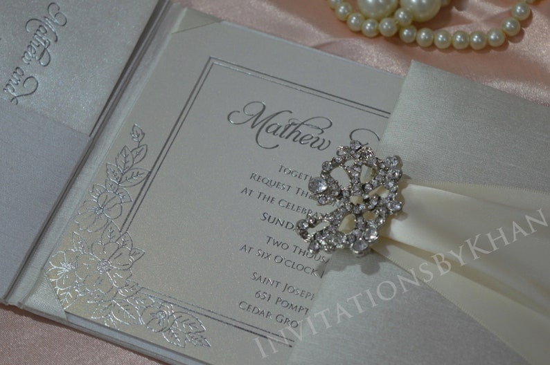 May include: A white wedding invitation with silver foil accents and a floral design. The invitation is tied with a white satin ribbon and a rhinestone brooch. The text on the invitation reads "Mathew and" and includes the wedding date and time.