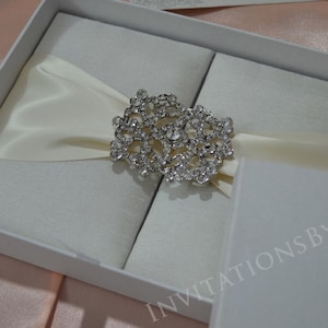 May include: A white satin box with a crystal embellishment, closed with a white satin ribbon. The box is labeled "Invitations by Khan".