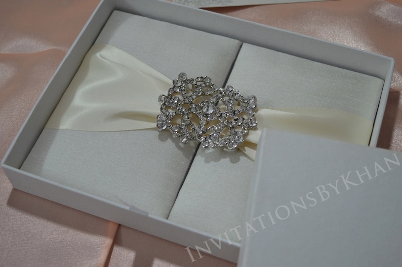 May include: A white satin box with a crystal embellishment, closed with a white satin ribbon. The box is labeled "Invitations by Khan".