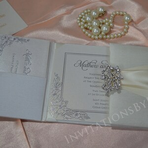 May include: A white wedding invitation with a satin ribbon and a rhinestone brooch. The invitation is folded and has a silver floral design on the front. The text on the invitation reads "Mathew and [name] Together request the pleasure of your company at the celebration of their marriage Sunday, [date] Two thousand [year] at six o'clock Saint Joseph's 631 Pompton Cedar Grove"