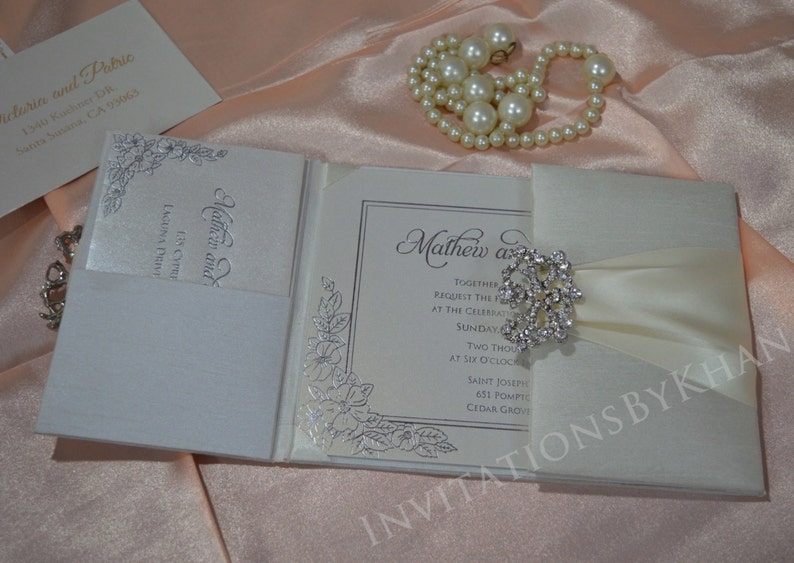 May include: A white wedding invitation with a satin ribbon and a rhinestone brooch. The invitation is folded and has a silver floral design on the front. The text on the invitation reads "Mathew and [name] Together request the pleasure of your company at the celebration of their marriage Sunday, [date] Two thousand [year] at six o'clock Saint Joseph's 631 Pompton Cedar Grove"