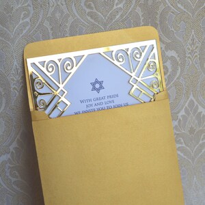 This is a beautiful, elegant Bar Mitzvah invitation featuring a striking gold laser-cut mirror acrylic frame with an intricate Art Deco style. The gold geometric swirls surround a white cardstock center printed with navy blue text and a Star of David at the top.

The combination of the formal serif typography for the details and the flowing script for Jacob's name creates a sophisticated and modern look.