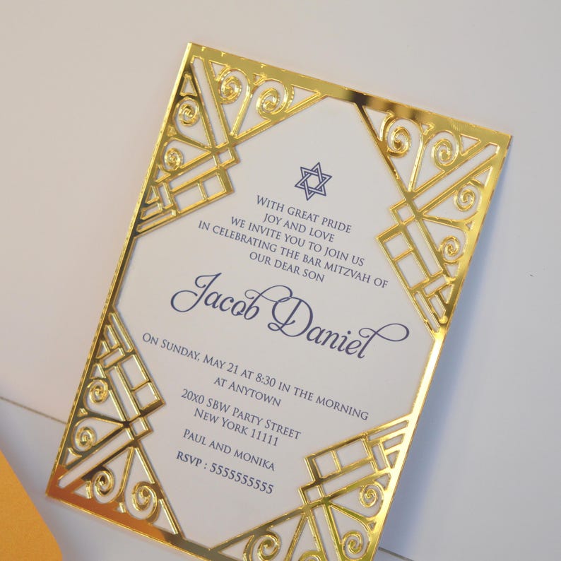 This is a beautiful, elegant Bar Mitzvah invitation featuring a striking gold laser-cut mirror acrylic frame with an intricate Art Deco style. The gold geometric swirls surround a white cardstock center printed with navy blue text and a Star of David at the top.

The combination of the formal serif typography for the details and the flowing script for Jacob's name creates a sophisticated and modern look.