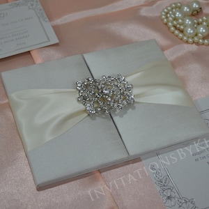 May include: A white satin wedding invitation box with a crystal brooch and a white satin ribbon. The box is closed and has a textured surface. The invitation box is on a pink satin background.