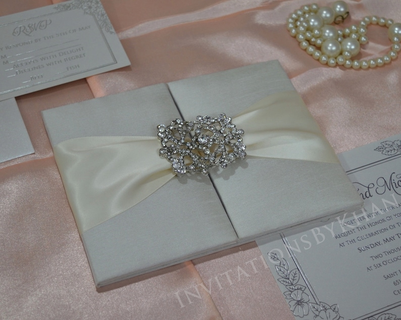 May include: A white satin wedding invitation box with a crystal brooch and a white satin ribbon. The box is closed and has a textured surface. The invitation box is on a pink satin background.