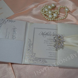 May include: A white wedding invitation with a satin ribbon and a rhinestone brooch. The invitation is folded and has a floral design on the front. The text on the invitation reads "Matthew and [name] Together request the pleasure of your company at the celebration of their marriage Sunday, [date] Two thousand [year] at six o'clock Saint Joseph's [address] Cedar Grove, [state]"