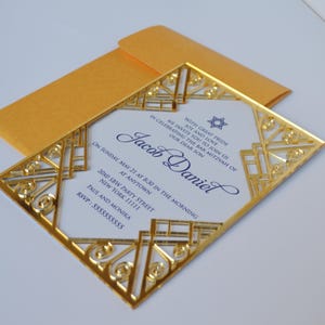 This is a beautiful, elegant Bar Mitzvah invitation featuring a striking gold laser-cut mirror acrylic frame with an intricate Art Deco style. The gold geometric swirls surround a white cardstock center printed with navy blue text and a Star of David at the top.

The combination of the formal serif typography for the details and the flowing script for Jacob's name creates a sophisticated and modern look.