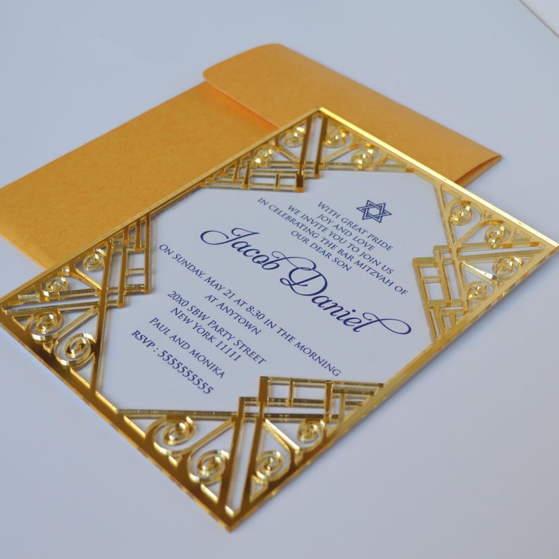 This is a beautiful, elegant Bar Mitzvah invitation featuring a striking gold laser-cut mirror acrylic frame with an intricate Art Deco style. The gold geometric swirls surround a white cardstock center printed with navy blue text and a Star of David at the top.

The combination of the formal serif typography for the details and the flowing script for Jacob's name creates a sophisticated and modern look.