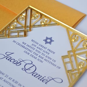 This is a beautiful, elegant Bar Mitzvah invitation featuring a striking gold laser-cut mirror acrylic frame with an intricate Art Deco style. The gold geometric swirls surround a white cardstock center printed with navy blue text and a Star of David at the top.

The combination of the formal serif typography for the details and the flowing script for Jacob's name creates a sophisticated and modern look.