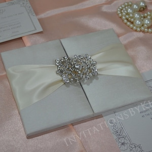 May include: A white wedding invitation with a satin ribbon and a rhinestone brooch. The invitation is on a pink background.