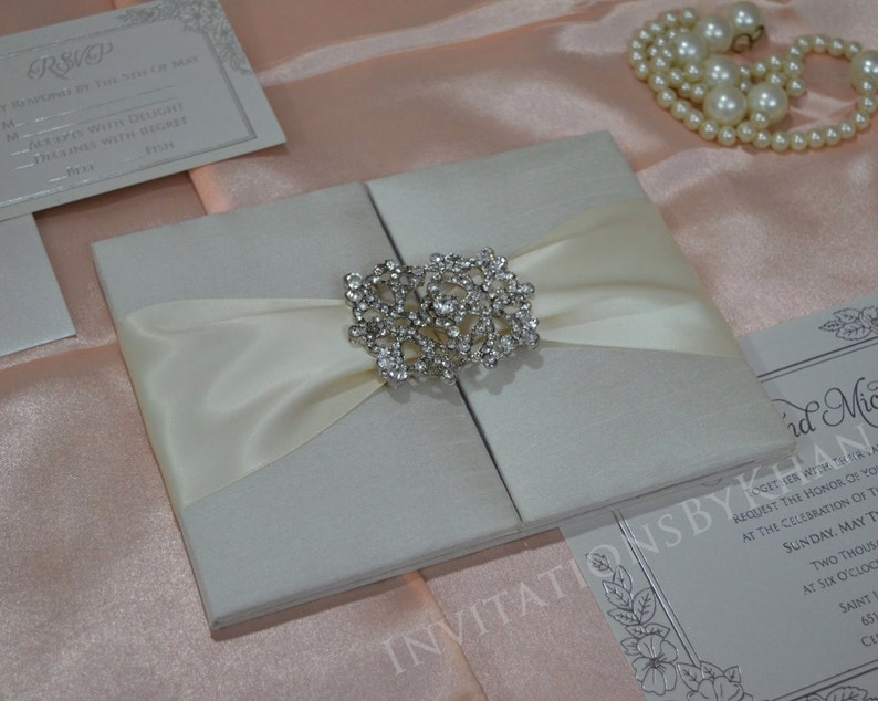 May include: A white wedding invitation with a satin ribbon and a rhinestone brooch. The invitation is on a pink background.