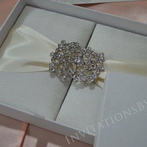 May include: A white satin wedding invitation box with a crystal embellishment. The box is closed and has the text "Invitations by Khan" printed on the bottom.