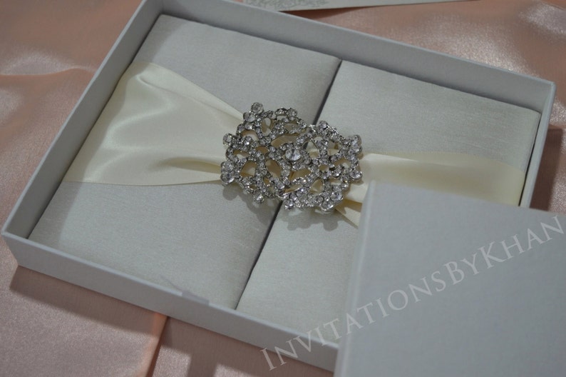 May include: A white satin wedding invitation box with a crystal embellishment. The box is closed and has the text "Invitations by Khan" printed on the bottom.