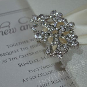 May include: A white satin ribbon with a silver rhinestone brooch. The ribbon is tied in a bow and the brooch is in the center of the bow. The text "INVITATIONSBYKHAN" is printed on the ribbon.
