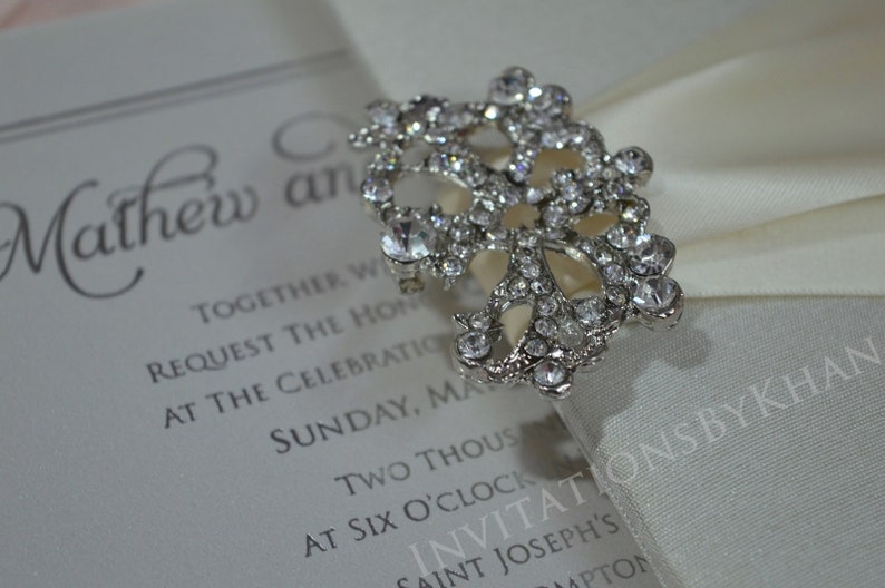 May include: A white satin ribbon with a silver rhinestone brooch. The ribbon is tied in a bow and the brooch is in the center of the bow. The text "INVITATIONSBYKHAN" is printed on the ribbon.