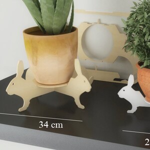 Peut inclure: Trois supports de plantes en bois en forme de lapins, chacun avec un pot et une plante de taille différente. Les supports mesurent 34 cm, 26 cm et 16 cm de large. Le plus grand support est en bois naturel, le support du milieu est blanc et le plus petit support est jaune.