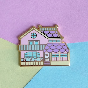 Könnte beinhalten: Eine rosa und lilafarbene Emaille-Anstecknadel mit einem Haus mit goldener Umrandung. Das Haus hat eine weiße Tür mit einem rosa Herzen und ein kleines Fenster mit einem blauen Rahmen. Das Dach ist lila mit goldener Verzierung und hat ein gewelltes Design.