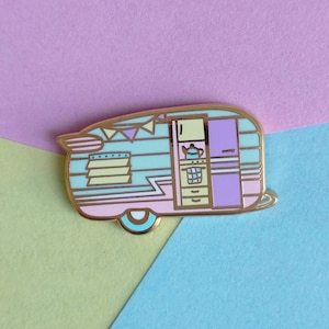 Könnte beinhalten: Eine pastellfarbene, rosa, blaue und grüne Emaille-Anstecknadel mit einem Vintage-Wohnwagen mit Fenster und Tür. Der Wohnwagen hat einen goldenen Umriss und eine kleine Flagge oben.