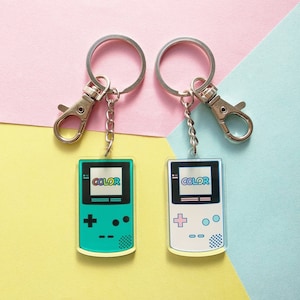 Puede incluir: Dos llaveros con forma de consola de videojuegos portátil. Los llaveros son de color verde y blanco con la palabra "COLOR" en la pantalla. Los llaveros están unidos a llaveros de plata.